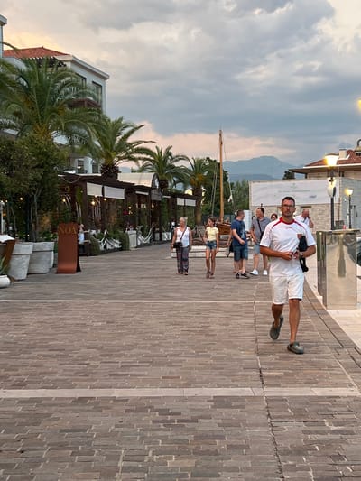 The image shows a scene on a paved walkway, likely a promenade, in Đenovići, Montenegro. The walkway is constructed with rows of brick-like paving stones. Several people are visible, appearing to be walking or standing.
In the foreground, a man is walking towards the camera, wearing glasses, a white polo shirt with red accents, and white shorts. He has a dark bag slung across his chest. Behind him, a few other individuals are walking in the same direction, including a man in a gray t-shirt and shorts, and another in a blue t-shirt and shorts. Further back, a woman in a yellow top and denim shorts is visible, as is another woman in a white top.
The setting includes buildings with balconies and awnings, some with white canopies. Palm trees are prominent in the scene, suggesting a warm climate. In the background, there are glimpses of hills or mountains under a cloudy sky, with the soft light indicating it is likely late afternoon or early evening. The overall mood appears relaxed and casual, consistent with a vacation or leisurely stroll in a coastal town.
There is visible text on a brown sign in the middle ground, appearing to read "VON" in white letters. The environment suggests a waterfront area, possibly near a harbor, with the presence of what looks like a mast from a sailboat.