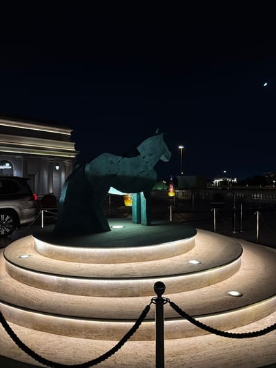 The image captures a nighttime scene in Doha, Qatar, featuring a modern stone sculpture and architectural elements. The sculpture, abstract in form and likely representing a horse, is situated to the left. Adjacent to the sculpture is a multi-tiered, curved architectural feature made of light-colored stone, possibly travertine or a similar material. Integrated lights illuminate each tier of the structure. A black rope is attached to a metal stanchion in the foreground. In the background, there's a hint of city lights and building facades. Part of a modern vehicle can be seen in the lower foreground, implying an urban setting. Above, the night sky is visible, with a faint crescent moon discernible. The setting suggests a public art installation in a well-lit, modern urban environment.