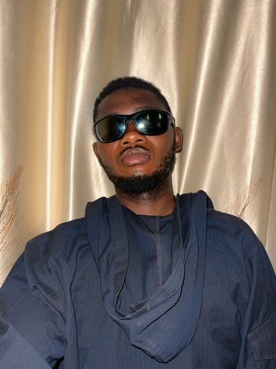 A man with dark skin, a black beard, and sunglasses faces forward. He wears a dark blue, hooded shirt. The background is a shiny, golden fabric draped vertically. The man's head is tilted to his right. The image is rotated to the left by 90 degrees.
