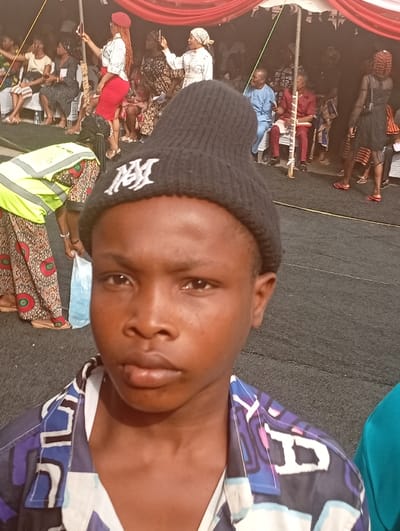 A young person wearing a black beanie with a white logo is in the foreground. The individual is wearing a patterned shirt. Behind them is a gathering of people under a tent. Many people are seated and some are taking photos with their phones. Someone in a red skirt and white top is standing and taking a photo, too. There is a person in a reflective vest holding a white plastic bag on the left side of the image.