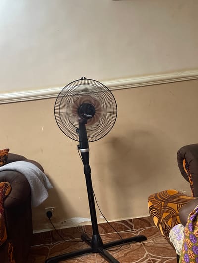This image captures an indoor scene in Narayi, Nigeria, depicting a living space.
A black Binatone pedestal fan is centrally positioned, operating with visible blade blur. The fan features a black circular grille and stands on a stable black cross-shaped base.
To the left, a dark brown upholstered sofa or armchair is partially visible, draped with a light grey towel. Beneath the sofa, a white wall outlet is visible on the light brown wall, with a black plug and cord connected, presumably powering the fan. The wall exhibits signs of wear, including peeling paint or plaster near the floor, revealing a lighter subsurface.
To the right, a portion of another upholstered furniture item is visible, featuring a distinct orange and dark brown swirling geometric pattern. A person's lower body, clothed in a vibrant purple, orange, and green floral-patterned fabric, is partially visible at the extreme right edge, adjacent to the patterned furniture.
The floor throughout the scene is covered with brown and tan patterned ceramic tiles. The light brown walls are topped with a white crown molding, separating them from the off-white ceiling. Minor scuff marks and discolorations are present on the walls and ceiling.