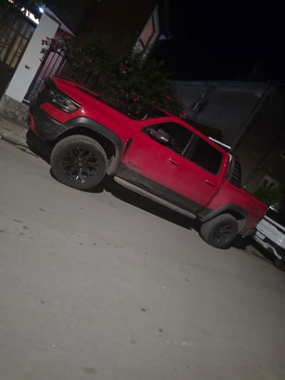 A red Ram TRX pickup truck is parked on a paved street at night in Targovishte, Bulgaria. The truck is angled, facing towards the left of the frame, with its front tires turned. The street is grey and appears to be illuminated by ambient light from nearby buildings and possibly streetlights. To the left of the truck, there is a white building with a fence and some greenery with red flowers. The truck has black wheels and black fender flares. The side of the truck displays the "RAM" logo. Further down the street, to the right of the truck, a white SUV is partially visible, also parked. The background consists of buildings that are dimly lit. The overall scene suggests a residential or semi-urban area at night.