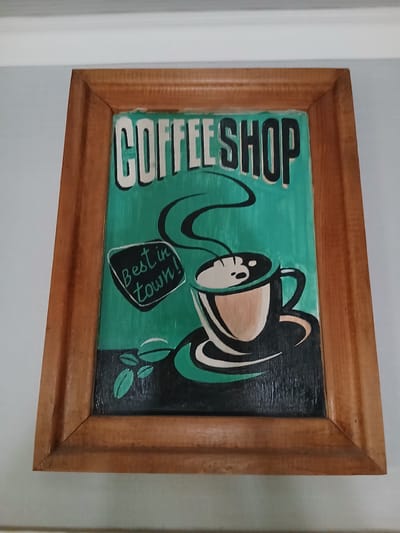 A framed painting hangs on a light-colored wall. The painting depicts a coffee shop scene with a turquoise background. At the top, the words "COFFEE SHOP" are written in bold, white, block letters with black outlines. Below this, a stylized coffee cup with steam rising from it is shown on a saucer. The cup is rendered in shades of white, beige, and black. To the left of the cup, a small black speech bubble contains white text that reads "Best in town!". At the bottom left of the painting, two green coffee beans are depicted. The painting is enclosed in a simple wooden frame. The image is a close-up, showing the upper portion of the painting and the frame, with a portion of a white ceiling visible at the top.