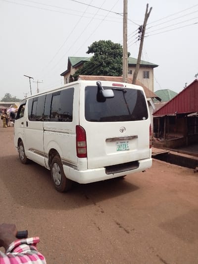 The image captures an outdoor scene on a hazy or overcast day, likely in a semi-urban or rural setting. The primary subject is a white Toyota Hiace passenger van, viewed from its rear-right side, positioned on a dirt or unpaved road. The van has tinted windows along its side and rear. A black external rearview mirror is mounted on the upper right side of its rear door. The rear features a Toyota emblem above a chrome strip, and below that, a white license plate with the registration "OKP 23LG". Faintly visible text above the license plate includes "NIGERIA" and what appears to be "RIVERS". The van shows signs of use, with dirt visible on its lower body panels and wheels.
In the background, several buildings are present. To the left, a light-colored, multi-story building with a green-tiled or green-painted gabled roof is visible, partially obscured by a large, leafy tree. To the right of the van, a simpler structure with a reddish-brown corrugated metal roof and wooden supports stands close to the road, featuring what looks like an open-front stall or shop. Another building with a light-colored facade is behind the green-roofed structure.
The sky is bright but appears uniformly white or light grey, indicating an overcast condition or haze. Numerous utility poles and power lines crisscross the upper portion of the image. One wooden utility pole with multiple cross arms and insulators is prominent on the right side, just behind the van. Another, possibly a street light pole, is visible further left.
On the far left side of the road, in the distance, a few people are faintly visible, seemingly gathered near what could be market stalls or a gathering point. In the immediate foreground, at the bottom left, the arm of a person wearing a red and white plaid shirt is visible, holding onto the black handlebar of what appears to be a bicycle or motorbike, suggesting the photo was taken from another moving vehicle or bicycle. The road itself is made of reddish-brown dirt or gravel, with some darker patches suggesting moisture or wear. An open drainage ditch or culvert runs alongside the road on the right side.