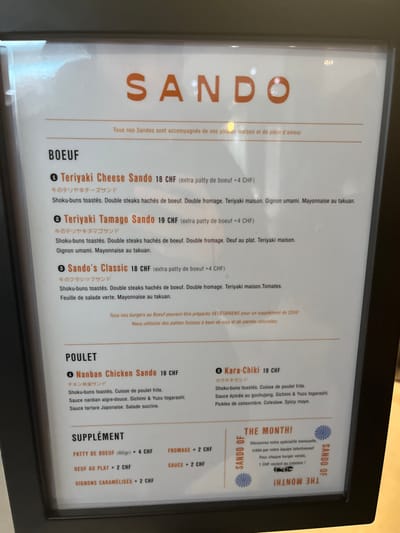 The image captures a menu board, likely illuminated, displaying food options. The setting appears to be indoors, possibly a restaurant or cafe, given the clear presentation of dishes and prices. No people are visible.
The menu is predominantly in French, with some Japanese katakana characters accompanying the dish names. The currency used is Swiss Francs (CHF), which aligns with the provided context of Genève, Switzerland.
The menu is organized into sections: "BOEUF" (Beef), "POULET" (Chicken), "SUPPLÉMENT" (Supplement), and a special "SANDO OF THE MONTH!".
Under "BOEUF", options include "Teriyaki Cheese Sando" (18 CHF), "Teriyaki Tamago Sando" (19 CHF), and "Sando's Classic" (18 CHF). These items feature toasted shoku-buns, double beef patties, various toppings such as cheese, teriyaki sauce, umami onions, takuan mayonnaise, fried egg, tomatoes, and green lettuce. An extra beef patty costs 4 CHF. A note indicates that any beef burger can be prepared vegetarian for a 2 CHF supplement, utilizing Swiss soy and plant-based patties.
The "POULET" section lists "Nanban Chicken Sando" (19 CHF) and "Kara-Chiki" (19 CHF). These sandwiches also use toasted shoku-buns and include fried chicken thigh, accompanied by different sauces (Nanban sweet-sour, Sichimi & Yuzu togarashi, Japanese tartare, gochujang, spicy mayo) and ingredients like sweet salad, cucumber pickles, and coleslaw.
The "SUPPLÉMENT" category offers additional items: an 80g beef patty (4 CHF), a fried egg (2 CHF), caramelized onions (2 CHF), cheese (2 CHF), and sauce (2 CHF).
The "SANDO OF THE MONTH!" section promotes a monthly special created by the team, stating that 1 CHF from each sale of this burger goes to the creator. The overall presentation is clean and functional, designed to inform customers about the offerings.