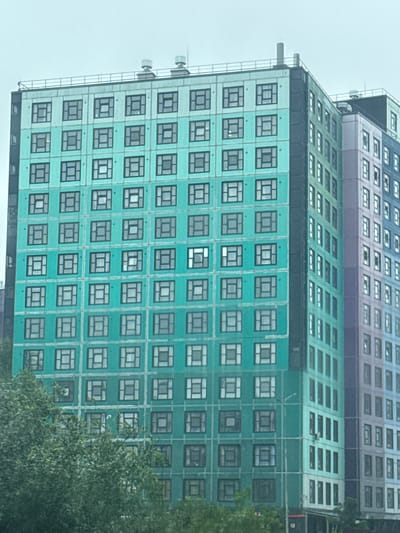 This media file is a medium-shot, vertically oriented photograph of a modern, multi-story building. The building appears to be constructed with a facade of teal-colored panels, interspersed with rows of windows. The windows are uniformly square or rectangular and framed in black. Some windows are dark, suggesting they are not illuminated from within, while others are lighter, possibly reflecting the overcast sky.

The building has a tiered structure, with the upper sections featuring different colors and window arrangements. The very top visible section is a darker, more muted color, possibly purple or dark gray, with rows of horizontally oriented rectangular windows. Below this, there is a section with a slightly lighter shade and a sloping roof-like structure covered in horizontal rows of windows. The teal section that forms the majority of the visible facade appears to be the main body of the building. The overall impression is one of urban architecture, likely in a city or suburban setting.

The sky is a uniform light gray, indicating an overcast or cloudy day. There is no discernible activity or people visible in the image. The focus is entirely on the architectural features of the building. To the right of the building, a dense cluster of green foliage from trees partially obscures the lower right portion of the facade, suggesting the building is situated in an area with some vegetation. There is a metal ladder or structure running vertically along the left side of the teal section of the building. No text is visible in the image. The location context provided is Murino, Russia.