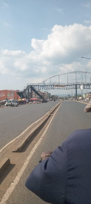 The image captures a daytime scene on a dual carriageway in Kisumu, Kenya. In the lower right foreground, a partial view of an individual's right arm and shoulder is visible, clad in a dark blue garment. A multi-beaded, light-colored bracelet adorns the wrist.

The asphalt road, divided by a low, light-brown concrete median with an integrated trench, extends into the background. To the left of the carriageway, a roadside market operates, featuring numerous individuals and stalls covered with colorful tarpaulin canopies in red, orange, and yellow. Two white vehicles, including a compact car, are positioned near the market. Behind the market, a multi-story building displays an orange facade with a large, partially legible advertisement.

A prominent dark-colored metal pedestrian footbridge spans horizontally across the entire width of the road in the mid-ground. This bridge exhibits a truss framework and includes access staircases descending to ground level on both sides of the carriageway. Streetlights are visible adjacent to the road and the footbridge structure.

The background features distant hills or mountains under a sky primarily composed of large white cumulus clouds on a light blue base.