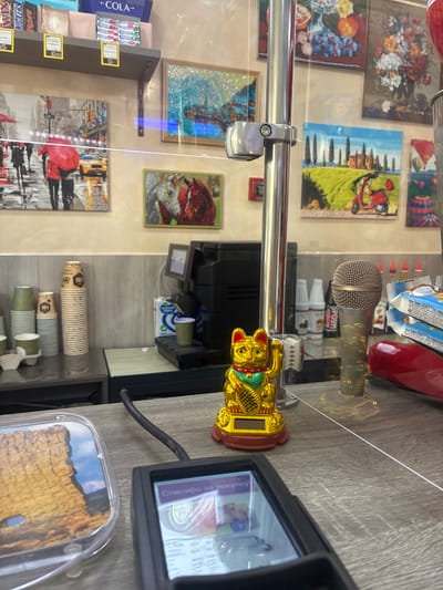 This is a close-up, eye-level, indoor photograph of a payment terminal, a Maneki-neko figurine, a microphone, and stacks of disposable cups on a counter. The payment terminal is in the foreground, with its screen displaying a message in Russian. To the left of the terminal is a clear plastic container with a picture of a rocky landscape and a blue sky inside. A golden Maneki-neko figurine, a traditional Japanese beckoning cat, is positioned behind the payment terminal. A microphone stands to the right of the figurine. In the background, several stacks of paper cups are visible, along with a coffee machine and various artworks on the wall. The overall setting appears to be a retail establishment. The artworks on the wall depict scenes of cityscapes, landscapes, and still lifes, suggestive of a tourist-oriented shop or cafe. The presence of the payment terminal and the coffee machine indicates a place of transaction and service. The specific location in Gelendzhik, Russia, cannot be determined from the visual information alone.