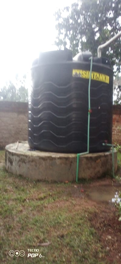 In Mahiakalo ward, Kenya, an outdoor scene depicts a large, black, corrugated plastic water storage tank prominently positioned on a circular concrete plinth. The tank bears a yellow label with the discernible text "SIM TANK". A white PVC pipe enters the tank from the top right, while a green PVC pipe extends vertically down the tank's side, featuring a red clip-like attachment, and then connects to additional green piping at the base, leading off to the right where a valve is visible. The ground surrounding the concrete plinth is covered in sparse green grass and appears damp, with visible patches of bare earth and minor puddling. Behind the tank, a low, red-brown brick wall is partially visible. The background consists of a bright, overcast sky and the silhouettes of trees or dense foliage. No individuals are present in the image, and no active processes or interactions are depicted beyond the static configuration of the water system. The bottom left corner of the image contains a "TECNO POP 6Go" watermark.