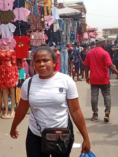 The image depicts a bustling open-air street market within the city of Asaba, Nigeria. In the foreground, a mid-adult woman of apparent Nigerian ethnicity walks towards the viewer, positioned slightly to the left of the frame's center. She wears a white patterned short-sleeved t-shirt with a dark emblem on the left chest, black trousers, and gold-colored hoop earrings. A dark crossbody bag, featuring a brown flap with the red text "MWTD," is slung over her left shoulder. Her right hand holds a light blue plastic bag, and her facial expression is neutral.

The midground and background are densely populated with various market stalls, showcasing an extensive array of new clothing items, including t-shirts (some with motifs like cherries), jeans, dresses, blouses, and skirts. These garments are displayed on hangers, racks, poles, and multiple mannequins, one of which exhibits a red floral-patterned dress to the left of the foreground woman. Overhead, dark canopies and a lighter-colored umbrella provide shade for the market area. Numerous individuals, including a man in a red t-shirt and grey baseball cap seen from behind on the right, navigate the crowded market space. The ground is a paved surface. Multi-story urban buildings are visible in the distant background under an overcast sky, establishing an active outdoor daytime commercial setting.