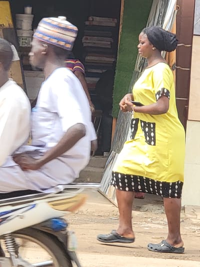In Maiburiji, Nigeria, a woman wearing a yellow dress with black and white trim stands on a concrete surface near what appears to be a storefront with stacked items inside. She is looking down at her hands. She wears black flats. Nearby, a man in a white outfit and a striped cap is bent over near a bicycle or motorcycle.
