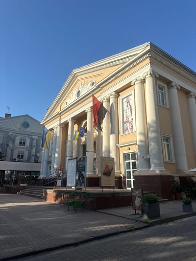 The image depicts the exterior of a building with classical architectural elements, including large columns and a triangular pediment. The building is painted a light beige color and features several windows. Several flags are visible, including Ukrainian flags and what appears to be the flag of the European Union. There are also signs in front of the building, one of which advertises "Dan Bakery" with an image of a croissant and another with an image of ice cream. The scene appears to be outdoors on a sunny day, as indicated by the clear blue sky and the shadows cast on the paved sidewalk. The building is situated on a street with a cobblestone pavement. The overall impression is one of a public or commercial building in an urban setting, likely in Ternopil, Ukraine, given the provided location context. There is a small bird flying in the sky. The image is taken from a low angle, looking upwards at the building.