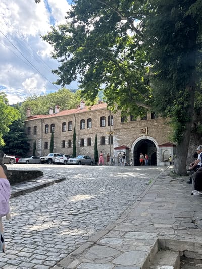 The image shows the entrance to the Bachkovo Monastery in Bachkovo, Bulgaria. The monastery is a large, stone building with a red tiled roof. The entrance is an arched gateway with an icon above it. Several people are entering and exiting the monastery. In front of the monastery, there is a cobblestone courtyard with several cars parked. Trees line the side of the courtyard. The sky is cloudy.