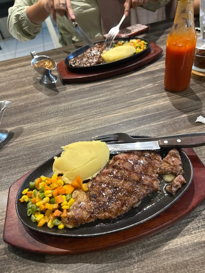 Two sizzling steak platters are displayed on a wooden table. The platters are black cast iron with wooden bases, a common way to serve hot dishes. The steak itself is thick-cut, appears well-seasoned, and is glistening with juices. It is served with a scoop of mashed potatoes and a colorful medley of mixed vegetables, including corn, carrots, and green beans.

In the foreground, the steak platter is prominently featured, with a fork and knife set beside it. The knife's handle is dark wood, and the fork is silver. A small metallic condiment holder, filled with a light brown sauce, sits to the left of the platter. To the left of this, a person's hands are visible, one holding a fork and the other a knife, indicating they are about to eat or are in the process of eating. The person is wearing a light green long-sleeved shirt.

Further back on the table, a second steak platter is partially visible, also featuring steak, mashed potatoes, and vegetables. Next to this platter is a glass bottle filled with a bright orange sauce, likely chili or tomato sauce, commonly found in Indonesian cuisine. The table has a rustic wooden texture, suggesting a casual dining setting. The lighting is warm, casting shadows and highlighting the glossy texture of the steak. The scene evokes the enjoyment of a hearty meal, likely in a restaurant in Makassar, Indonesia, given the location context. There is no visible text in the image.