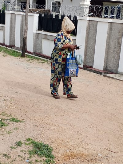 A woman stands on a sandy road in Adonkolo, Nigeria. She is wearing a patterned, multi-colored two-piece outfit and a light-colored head covering. She holds two cellphones in her hands and looks down at them. She carries a blue tote bag with the words "KEDI HEALTH" printed on it, along with a small white fan. The road is unpaved and sandy, with patches of vegetation. Behind her is a wall topped with barbed wire. A utility pole stands to the left of the wall.