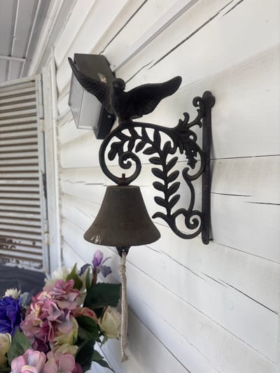 This is a close-up, overhead shot of a black, cast iron wind chime. The wind chime features a decorative design with a bird on the left and a bell on the right, suspended from a wrought iron bracket. The bracket is attached to a white, wood-paneled ceiling or overhang. A small, dark outdoor light fixture is positioned behind the bird. A thick, beige rope is tied through the top of the bell and extends to the right, where it is intertwined with a floral arrangement. The flowers are a mix of colors including pink, purple, white, and yellow, with green leaves. The scene appears to be outdoors, possibly on a porch or under an eave. The time of day is indeterminate due to the lighting, but the overall impression is one of rustic charm. The specific location is identified as Лотос Снт (марьино), Russia. There is no visible text in the image.
