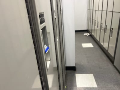 The image depicts a hallway with a row of lockers. One locker, numbered 101, is prominently featured on the left side of the frame, with a blue item visible inside its compartment. Further down the hallway, another set of lockers is visible, with a pair of white flip-flops or sandals on the floor in front of them. The floor is tiled, and there are squares of light on the floor, suggesting overhead lighting. The scene appears to be an indoor facility, possibly a gym, school, or workplace. This image was taken on the Burnaby campus of a Canadian university.
