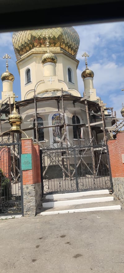 The image shows an exterior view of a church in Kolomak, Ukraine. The church has multiple golden domes and is under scaffolding, suggesting construction or renovation work. The building's facade is a light yellow color, with arched windows and crosses on top of the domes. In the foreground, there is a black wrought-iron gate with an ornate design, and a brick pillar with a green informational sign. White steps lead up to the gate. The ground in front of the gate is asphalt, with some dark spots. The sky is blue with scattered clouds. The overall scene captures a religious building in a state of upkeep or development.