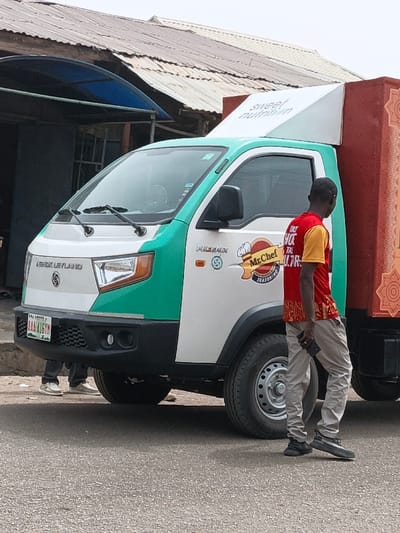 A daytime street scene in Gwagwarwa, Nigeria, depicts a white and green Ashok Leyland Phoenix light commercial truck parked on an asphalt road. The truck's front features the 'Ashok Leyland' logo, headlights, and a black bumper. A Nigerian license plate, registered in Lagos State as "AAA-416YM," is affixed to the front. The side of the truck's cab displays a "Mr. Chef Seasoning" logo, while a white angled panel above the cab reads "Sweet Nutrition." The cargo box behind the cab is reddish-brown with an intricate, darker patterned design. A male subject, walking from right to left, is positioned next to the truck. He wears a red and yellow short-sleeved shirt, light-colored trousers, and dark athletic shoes, holding a dark-colored smartphone in his right hand. The lower legs and feet of another individual, wearing light-colored sneakers, are partially visible beneath the front section of the truck. In the background, structures with corrugated metal roofs (one silvery-grey, another brownish-red) and a blue awning are visible.