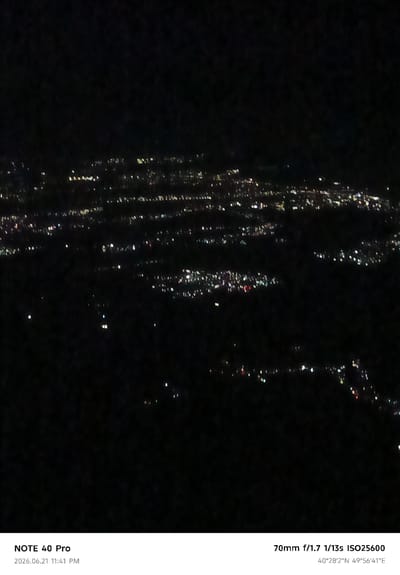 The image is a night-time aerial view of a city, likely Dushanbe, Tajikistan, illuminated by countless small lights from buildings and streetlights spread across a dark landscape. The lights are out of focus, appearing as blurry dots and streaks, suggesting a long exposure or significant distance from the camera. The composition is dominated by darkness with the city lights forming a distinct pattern in the mid-ground and extending towards the horizon. The lower portion of the image displays text overlays indicating camera settings and the date and time of the photograph: "NOTE 40 Pro", "2026.06.21 11:41 PM", "70mm f/1.7 1/13s ISO25600", and coordinates "40°28'2"N 49°56'41"E". There are no visible people or specific objects, only the abstract representation of a city at night. The overall impression is one of a vast, urban expanse seen from above in low light conditions.