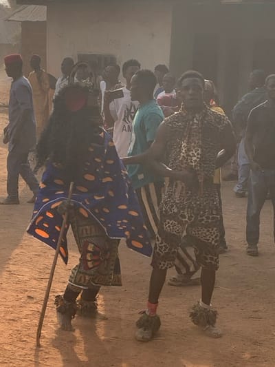 A cultural scene unfolds outdoors, with a person adorned in a large, dark mask and vibrant blue, orange, and yellow patterned fabric, carrying a wooden staff. Behind them, a person in a leopard-print outfit gestures actively. A crowd of onlookers gathers in the background, some holding phones, amidst a dusty setting with low buildings and a hazy atmosphere. The scene appears to capture a traditional performance or ceremony.
