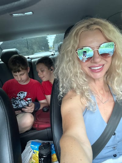 The image shows a woman with blonde, curly hair and sunglasses taking a selfie in a car. She is smiling and wearing a light blue top. Her sunglasses reflect a scene of trees and buildings, possibly indicating an outdoor environment. In the back seat of the car, two young boys are visible. One boy is leaning on the other, and both appear to be sleeping or resting. They are both wearing red t-shirts. One of the boys has a blue band on his arm.

Visible objects in the car include a black water bottle and some snack bags. The scene appears to be set during the daytime, with natural light coming from outside. The presence of a car suggests they are traveling. The overall emotion conveyed by the woman is happy and relaxed, while the boys seem peaceful in their slumber. There is no visible text in the image. The location is Goryachy Klyuch, Russia.
