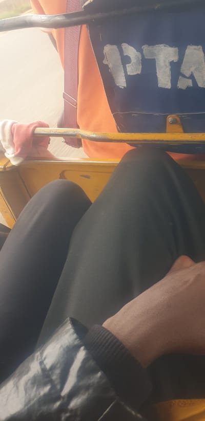 The image shows a close-up view of the interior of what appears to be a yellow rickshaw in Gwong, Nigeria. The dominant subjects are parts of two people. One person is wearing black pants, and their hand and forearm are visible on the right side of the frame. The skin on their forearm shows prominent veins. They are wearing a black jacket or shirt with a ribbed cuff.
On the left side of the rickshaw, an orange fabric is visible, likely part of the seating or a covering. A blue bag or piece of fabric with white lettering is also present. The visible text reads "PTA" with some faded or smudged letters before it.
A yellow metal bar, part of the rickshaw's structure, runs vertically through the middle of the image. A hand wearing a red and white glove is gripping this bar, suggesting the person is a passenger holding on.
The lighting suggests it is daytime, with soft, diffused light. There are no clear indicators of weather conditions. The overall impression is a snapshot of daily life and transportation within a Nigerian context. The orientation of the image is also rotated 90 degrees clockwise.