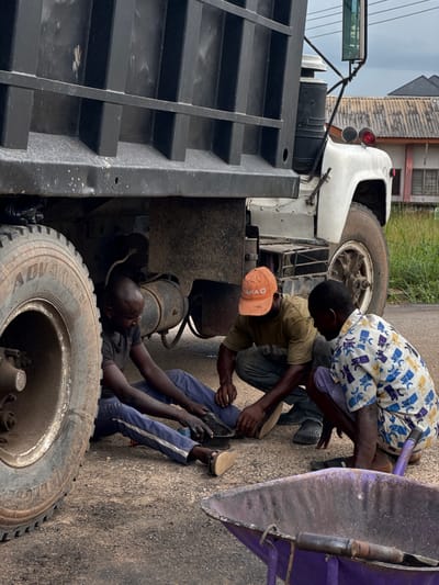 The image captures three men engaged in the repair of a large truck on a paved surface, likely a roadside in Ihiagwa, Nigeria. The scene unfolds during the daytime under an overcast sky.
In the foreground, a large truck tire is prominent, with the words "ADVANCE" and "GLOBAL" visible on its sidewall. One man, dressed in a dark t-shirt and blue pants with white stripes, is lying on his back underneath the truck, actively working on its components.
Two other men are crouching beside the truck. One wears a white and blue patterned shirt, while the other has a light brown t-shirt and an orange baseball cap with "MIA" partially visible. They appear focused on their tasks, with their hands busy near a purple metal object that might be part of the truck's machinery.
The truck itself appears to be a dump truck, with its large black bed partially visible on the left and a white cab section seen towards the top. In the background, simple buildings and some greenery are visible, characteristic of a semi-urban or rural setting. The overall image conveys a dedicated, hands-on repair effort.
