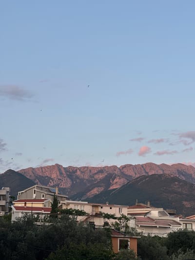The image depicts a scenic view of mountains and buildings, possibly in Šušanj, Montenegro. The sky is a light blue with scattered pinkish clouds, suggesting it's either sunrise or sunset. The mountains in the background are rugged and rocky, with a darker, forested slope below. In the foreground, there are several buildings with white and red roofs, appearing to be residential or multi-story structures. Green trees and foliage are interspersed among the buildings and along the right edge of the frame. There are at least three small dark shapes in the sky that could be birds in flight. The overall mood is peaceful and serene, characteristic of a natural landscape at dawn or dusk.