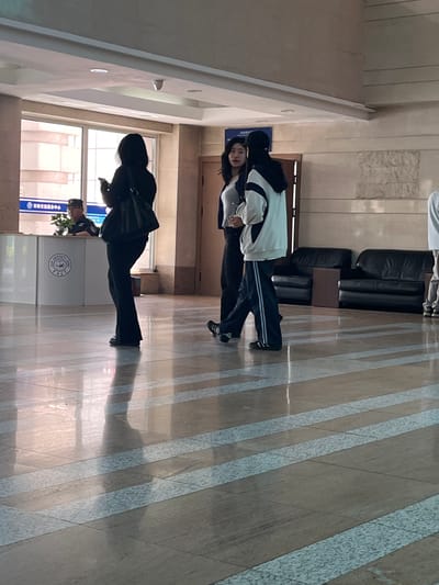 The image captures an indoor scene, likely a university or administrative building lobby in Changchun City, China. In the foreground, a polished floor with a distinctive pattern of beige tiles and light blue-grey strips reflects the overhead lighting. A person on the left is shown from the knees down, wearing dark, flared pants and dark shoes, holding a dark bag over their left shoulder and a mobile phone in their right hand. They are facing away from the camera and to the left.
Further into the scene, two individuals are standing closer together, facing each other in conversation. The person on the left, partially obscured, has dark hair and is wearing a light-colored top and dark pants. The person on the right wears a black baseball cap, a white and grey hooded jacket over a dark top, and dark pants with white stripes. Their posture suggests an ongoing interaction.
Behind them, a white reception desk with a circular blue and white logo is visible. A uniformed individual, likely a security guard, is seated behind the desk. In the background, a wall with a stone-like texture is visible, along with two dark leather sofas and a small wooden table. Large windows with a building facade visible through them are to the left of the reception desk, and a door is to the right of the two conversing individuals. The overall environment appears well-lit and clean, typical of a modern public building.