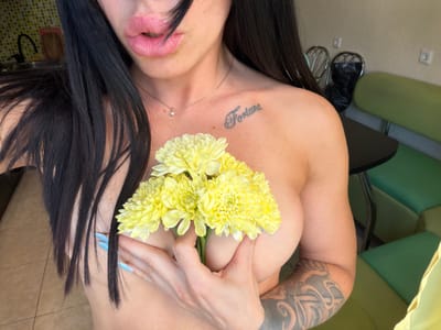 The image shows a close-up of a woman holding a bouquet of yellow flowers in front of her chest. She has long, dark hair and is wearing a necklace. There is a tattoo on her collarbone that says "Fortune". Her fingernails are painted light blue. In the background, there are green chairs and a table, suggesting an indoor setting. The floor appears to be tiled. The image does not contain any identifiable landmarks or details that would place it specifically in the city of Ufa, Russia.