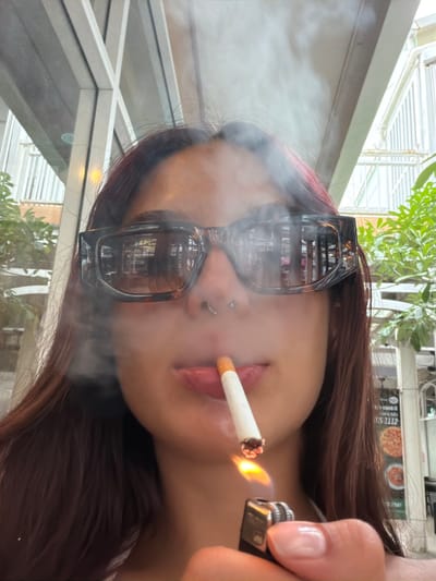 The image shows a close-up of a young woman with long, dark hair, wearing oversized, tortoiseshell sunglasses and a nose ring. She is holding a lit cigarette to her lips, with smoke emanating from it. Her right hand is visible, holding a black lighter with a flame igniting the cigarette. The background appears to be an outdoor seating area, possibly a cafe or restaurant, with glimpses of greenery and architectural elements suggesting an urban environment. There is a sign in the background that seems to advertise food, with images of pizzas. The overall lighting suggests it is daytime. The location is identified as Choeng Thale, Thailand.