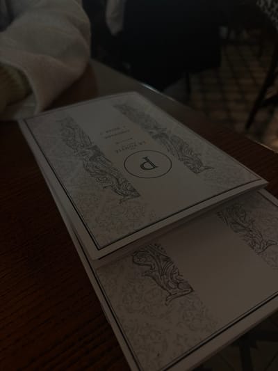 An open menu sits on a wooden table. The cover features a decorative design with a large letter "P" encircled. Below the "P" is text, including the words "LE PONTIE", "LIMONADE", and "RIVIERA". The open pages are mostly blank, framed by similar decorative elements as the cover. A white fabric is visible at the bottom left corner of the frame.