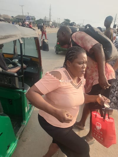 A woman in a pink top and black pants is standing on a street near a green motorized tricycle. She holds tissues in one hand. People pass behind her, with a red shopping bag visible. A man with a black backpack is partially visible in the background. The tricycle is parked on the side of a paved road, and a driver is seen sitting inside. Power poles and buildings are visible in the distance under a bright sky.