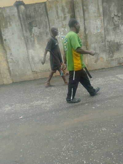 Two males are depicted walking leftward on a paved road surface adjacent to a weathered concrete wall in Agani, Nigeria.
The individual in the foreground (right) is a male wearing a short-sleeved green and yellow t-shirt with partial text, black athletic pants with a white vertical stripe, and dark Crocs-style sandals. A dark strap or belt hangs from his right hip.
The individual slightly behind and to the left is a slender male wearing a dark short-sleeved shirt, dark shorts, and dark flip-flops. He is carrying a small, multi-colored object (red, blue, metallic) with a chain in his right hand. Both individuals appear to be moving in the same direction.
The ground is a dark grey asphalt or concrete surface, unevenly covered with light brown dust and small aggregates. A small piece of red and white debris is visible on the road surface in the lower-left quadrant. The background consists of a tall, light-colored concrete wall, segmented vertically, with a circular embossed design in the upper-left portion and visible remnants of faded posters or paper adhered to its surface. The wall exhibits signs of weathering and discoloration. The scene is illuminated by ambient daylight.