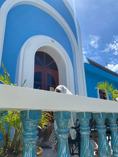 The image shows the exterior of a bright blue church with white arches against a clear blue sky with scattered clouds. A cross is visible on the roof. In the foreground, a white railing with blue decorative balusters separates the viewer from the church. Lush green palm fronds and other foliage are visible behind the railing. A person wearing a white head covering and striped clothing is partially visible behind the railing, leaning forward and looking down. The architecture suggests a location in Ko Samui, Thailand, known for its vibrant blue and white structures, often seen in religious buildings and traditional island architecture.