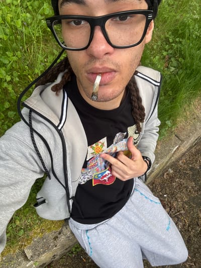 A man lies on his back in a grassy area, wearing black-framed glasses, a black t-shirt with a colorful graphic design, a grey zip-up hoodie with white stripes on the sleeves, and grey sweatpants. He has dark dreadlocks. A lit cigarette or joint is in his mouth. He is lying on grass alongside a concrete curb. The photograph appears to have been taken at ground level, angled upwards. The location could not be determined from this image alone.