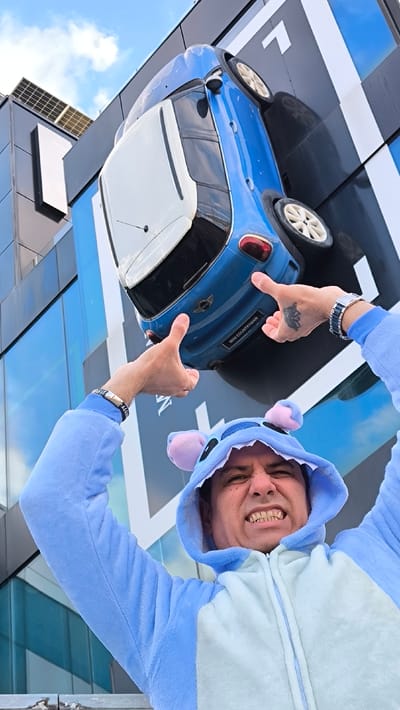 A man in a blue Stitch costume poses with outstretched arms, seemingly holding up a blue Mini Countryman mounted on a building. He wears a watch and bracelet on one wrist and has a tattoo on the other hand. The building features a modern design with glass panels and a black and blue facade. Solar panels are visible on the roof. The sky is partly cloudy.