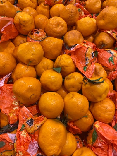 The image shows a close-up, overhead view of a pile of tangerines or small oranges. The fruits are various shades of orange and have textured skin. Some are individually wrapped in crinkled orange paper with Chinese characters and some English text that reads "THE BEST SW..." and "NESS!". The tangerines are densely packed, obscuring the surface they are resting on. The scene appears to be a market stall in North Jakarta, Indonesia, possibly at a traditional market or a fruit stand, given the common practice of selling fruits in such wrappers, especially during festive seasons. There are no people visible in the image.