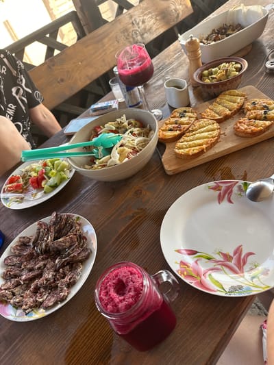 A high-angle, outdoor shot captures a wooden table laden with food and drinks. The scene is set in a rustic outdoor dining area, possibly a patio or garden, suggested by the wooden beams and lattice structure visible in the background. The lighting indicates it is daytime, with bright sunlight casting shadows.
Dominating the table are several dishes of food. A platter of sliced cured sausages sits on a white oval dish, while grilled bread slices with a golden crust are arranged on a wooden board. A large bowl contains a salad with what appears to be cabbage and tomatoes, served with bright green salad tongs. Another smaller oval dish holds a portion of salad with cucumbers, tomatoes, and pasta or noodles.
To the left, a white oblong dish contains what looks like sauteed mushrooms or vegetables, with a wooden salt or pepper grinder nearby. A small white pitcher holds a pale liquid, likely a sauce or dressing. A glass jar filled with a vibrant pink, frothy drink, possibly a smoothie or juice, is positioned near the plate of sausages.
A white plate with a floral pattern, featuring pink lilies and green foliage, is placed to the right, with a silver spoon resting on it. The overall impression is of a casual meal or gathering, with a focus on fresh and hearty food. A person's arm and torso are visible at the bottom left of the frame, wearing a black shirt with white print, suggesting they are seated at the table. The mood appears relaxed and convivial. No visible text is discernible in the image.