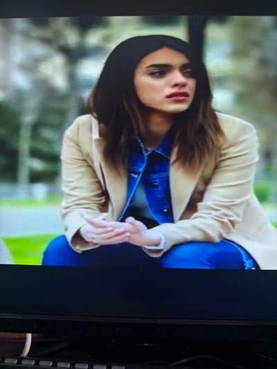 The image displays a close-up of a person with long, dark hair, wearing a beige blazer over a blue denim jacket and blue jeans. The person is leaning forward, with their head tilted and eyes partially closed, suggesting a pensive or tired expression. Their hands are clasped together in front of them. The background is blurred and appears to be an outdoor setting with green and light-colored foliage, possibly indicating a park or garden. The lighting suggests it could be daytime. There is no visible text in the image.