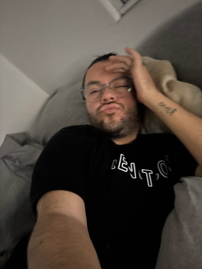 A man lies on a grey couch, his face obscured by his hand. He wears glasses and a black t-shirt with white lettering. His arm bears a tattoo. A beige blanket is near his head. The location is suggested as San Juan Cuautlancingo, Mexico, but no details in the image confirm this.