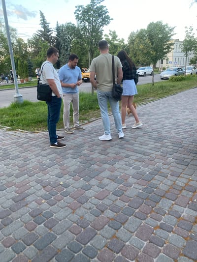 The image depicts a group of four individuals standing on a paved walkway in Poltava, Ukraine. The walkway is constructed of interlocking, multi-colored cobblestones. To the left of the frame, two men are engaged in conversation. One man, on the left, wears a white t-shirt and dark jeans, with a black messenger bag slung across his chest. The other man, to his right, is dressed in a light blue polo shirt and beige trousers. Beyond them, a yellow taxi is visible.
Further to the right, a man in a light olive-colored t-shirt and light blue jeans stands facing away from the camera. Beside him, a woman in a dark plaid jacket and a short, light blue skirt with a floral pattern is also facing away. A small, light brown dog is walking behind the woman. In the background, several cars are parked along a street, and trees line the area. A building with multiple windows is visible behind the trees on the right side of the image. The sky appears overcast.