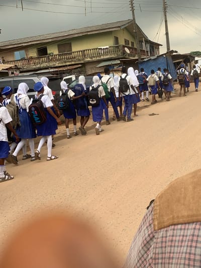 In Shagamu, Nigeria, a group of schoolchildren in blue and white uniforms are walking along a dusty road. They are wearing white headscarves and carrying backpacks. The children are walking in a line, with some children in the front and others following behind. In the background, there are buildings and a street. The sky is cloudy. The image appears to have been taken from a low angle, possibly from a vehicle or from someone walking in the same direction as the children.