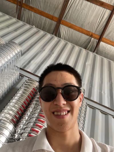 A young male, wearing round, dark-lensed sunglasses and a white collared shirt, smiles at the camera. He has short, dark hair. The background consists of corrugated metal walls and a ceiling made of metal sheeting and wooden beams. Several large, cylindrical, metallic ducts with red caps are visible, running vertically and diagonally. The image appears to be taken in a warehouse or industrial building. This location is specified as being in the village of Kenen Azirbayev, Kazakhstan.