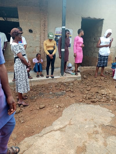 A group of people, mostly young women, stand near a dilapidated building. One young girl sits on the ground, head in hands. Some of the others stand in a row, casually dressed. The ground is uneven and broken. A pole stands in front of the building, partially obscuring a sign. The overall atmosphere is somber, and the area appears rundown.