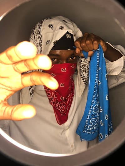 The image depicts an individual wearing bandanas of different colors—red, blue, black, and white. A white shirt is also visible. The person's face is partially covered with the red bandana, revealing only their eyes. One hand is clenched, holding the blue bandana, while the other is extended towards the viewer. The shot is taken from an unusual angle, possibly from below, and is framed by a circular light.