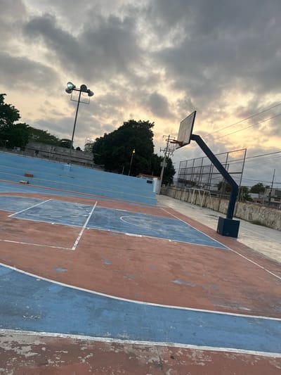 The image shows a public basketball court in Mérida, Mexico. The court surface is painted in blue and orange, with visible wear and tear. Two basketball hoops are present. One is closer to the camera and appears newer. A metal fence surrounds the court, with some greenery and trees visible beyond. A streetlight is on the left side of the frame. The sky is cloudy with a yellowish hue. The image is oriented vertically.
