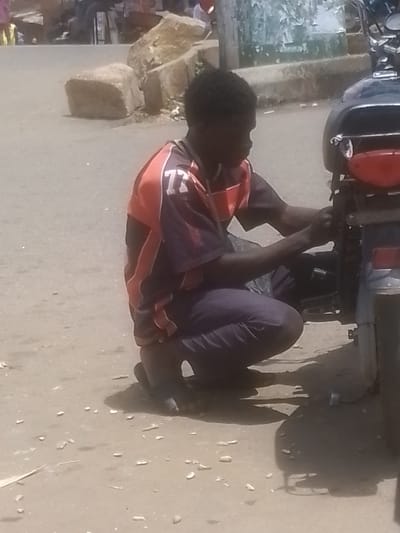 In Gwong, Nigeria, a person is seen crouching down and working on a motorcycle. The person is wearing a dark blue tracksuit with red and white stripes on the sleeves and a number "77" printed on the upper left chest area of the shirt. The individual's hair is dark and curly, and they appear to be focused on the task at hand. The motorcycle is dark in color and has a red taillight. The scene appears to be outdoors, with a dusty, unpaved ground. In the background, there are large rocks and a wall with some peeling posters or paint. The lighting suggests it is daytime, possibly late morning or early afternoon, with strong shadows. The overall atmosphere seems casual and practical, perhaps indicating a roadside repair or maintenance activity. No discernible emotions are evident due to the distance and resolution.