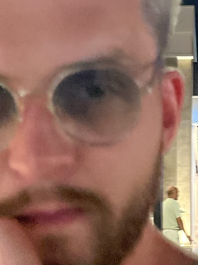 This close-up, distorted image features a person's face and eye in the foreground, appearing to be looking into a mirror or at a camera lens. The eye is dark and somewhat obscured by the camera's close proximity and focus. A reddish object, possibly a finger or a part of clothing, is visible in the upper left portion of the frame. In the background, a blurred scene shows another person lying down on what appears to be a light green surface, possibly a bed or lounge chair. This background figure is wearing a light-colored shirt and is facing away from the camera. The setting appears to be indoors, with soft lighting suggesting it could be daytime or evening. The overall image has a hazy, low-resolution quality. No text is visible.